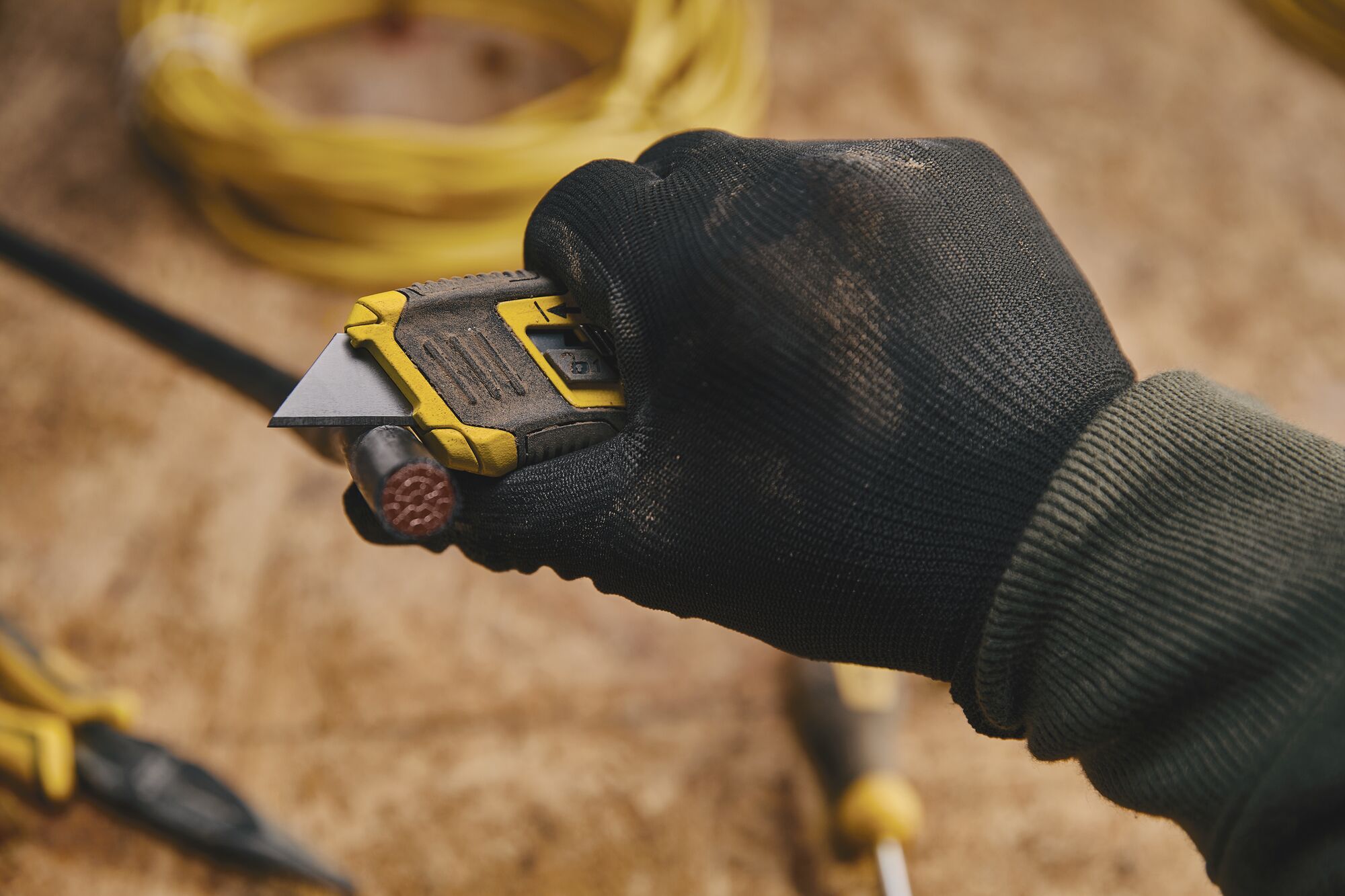 STANLEY STHT10432 utility knife in gloved hand cutting a cable, close-up with tools visible in background.