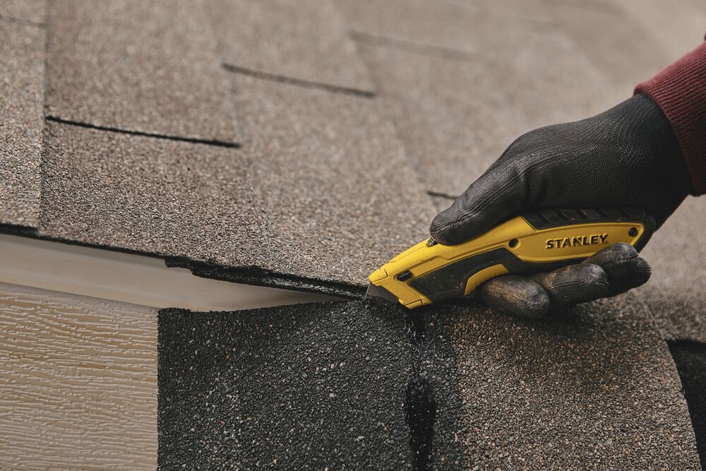 STANLEY STHT10479 utility knife cutting roofing shingles, close-up of gloved hand holding yellow and black tool.