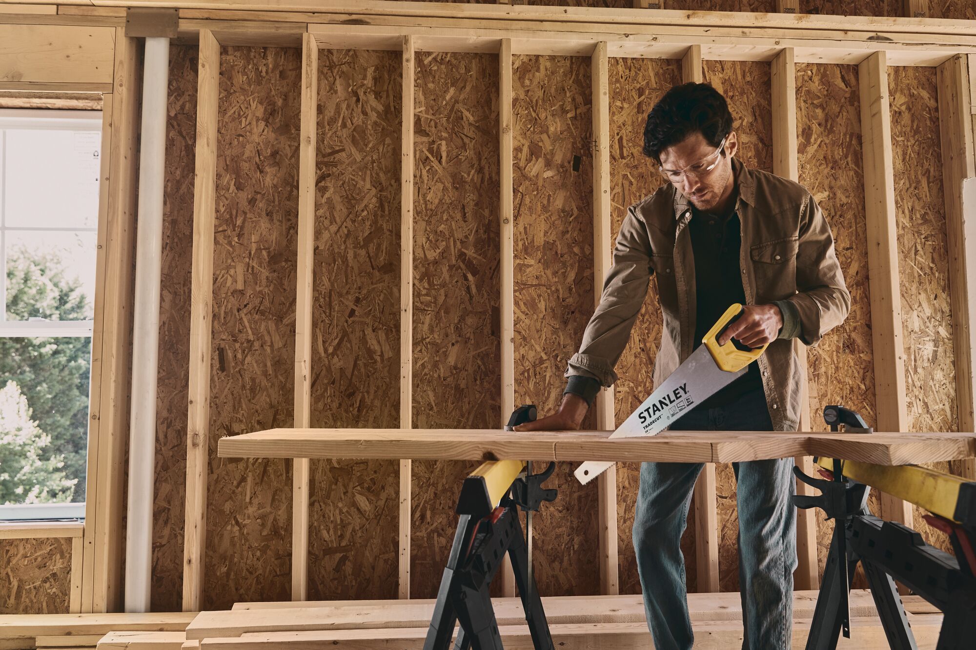 STANLEY STHT20350 hand saw cutting wood on sawhorses inside a framed construction area.
