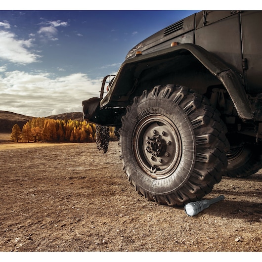 STANLEY TL10PS flashlight under large off-road vehicle tire on dirt with autumn trees in background.